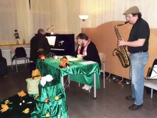 Foto: Ich sitze an einem reich geschmückten Lese-Tisch. Im Vordergrund Ulf am Saxophon, im Hintergrund Stephan am Piano.