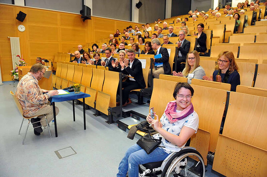 Foto: Ich sitze am unteren Ende des Hörsaales an einem kleinen Tisch und lese meine Texte vor. In den Rängen sitzen zahlreiche Zuschauer.
