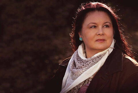 Foto: Suta Wanji mit rötlichen Haaren in brauner Jacke hat ein helles Halstuch um und schaut nach links aus dem Bild heraus.