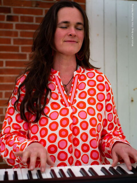 Marie-Lou sitzt mit einer Bluse mit rosaroten Kringeln an einem Piano und spielt Musik.