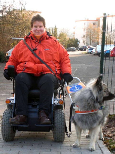 Foto: Susanne in knallrotem Anorak im E-Rolli und Filou im Führhundgeschirr links neben ihr.