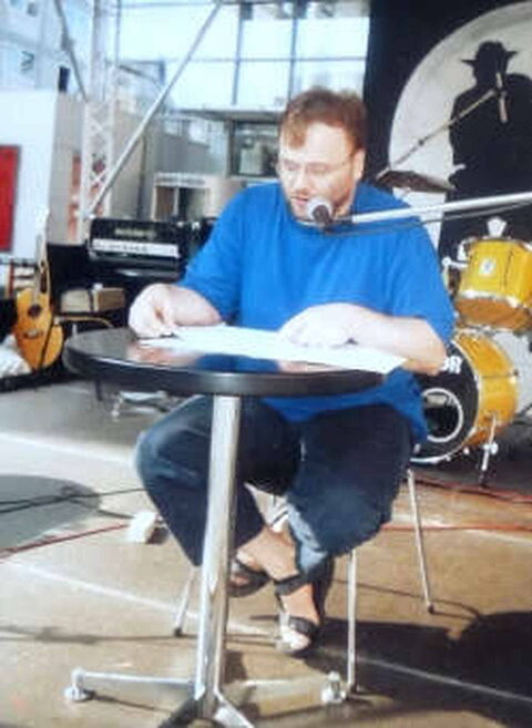 Foto: Ich sitze in blauen Klamotten auf einer Bühne an einem runden Tisch und lese einen meiner Texte vor.