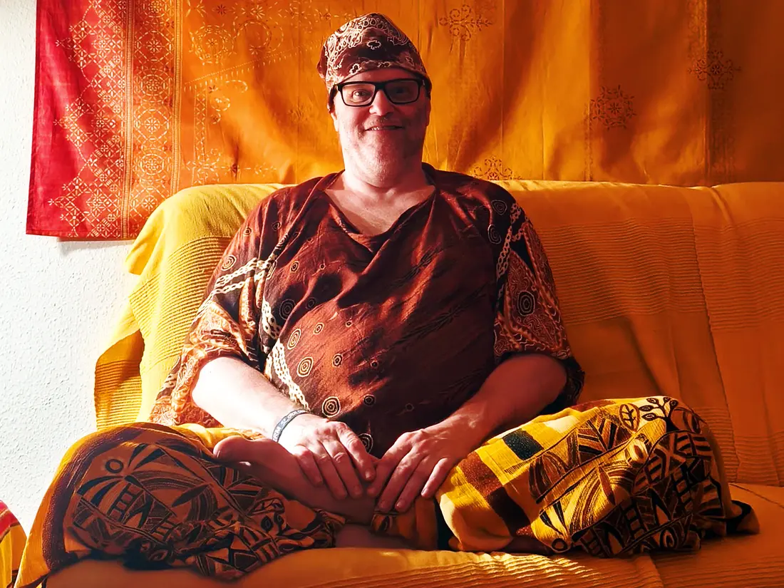 Foto: Ich sitze mit einer gelben Wickelhose, einem braunen Oberhemd und braun verziertem Kopftuch  auf meinem Sofa im Snoozleraum und schaue in die Kamera.