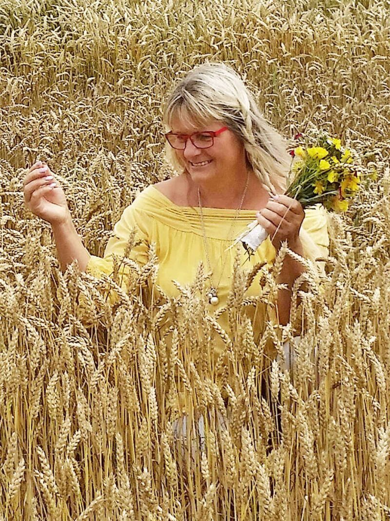 Foto: Brigitte mit einem gelben Hemdi nmitten eines wunderschönen Weizen-Feldes. In der linken Hand ein Blumenstrauß.