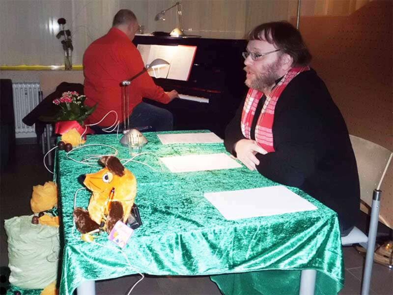 Foto: Meine Schreibmaus und ich sitzen an einem reich dekorierten Tisch und lesen Texte vor. Im Hintergund stzt Stefan am Klavier.