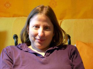 Foto: Evelyn in einem lila Hemd mit ihrem unwiderstehlichen Lächeln vor meinem gelben Sofa.
