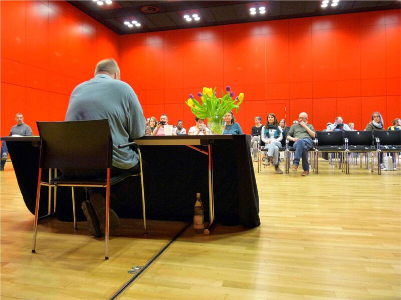 Foto: Ich sitze im Vordergrund an einem geschmückten Tisch, im Hintergund mein Publikum in einem großen Saal.