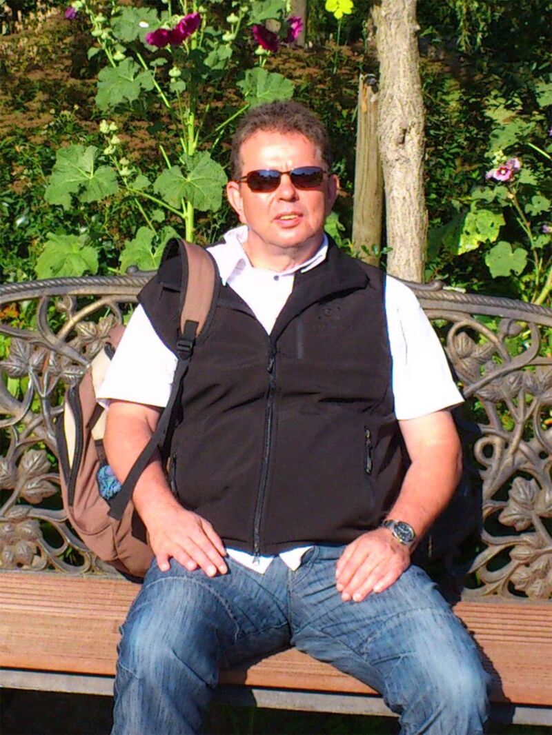 Foto: Joachim sitzt mit einem Rucksack über der rechten Schulter auf einer Parkbank in der Sonne, umgeben von Grün.