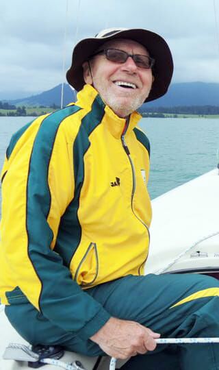 Foto: Jürgen sitzt im grün-gelben Trainingsanzug auf der Bootsreeling und segelt.