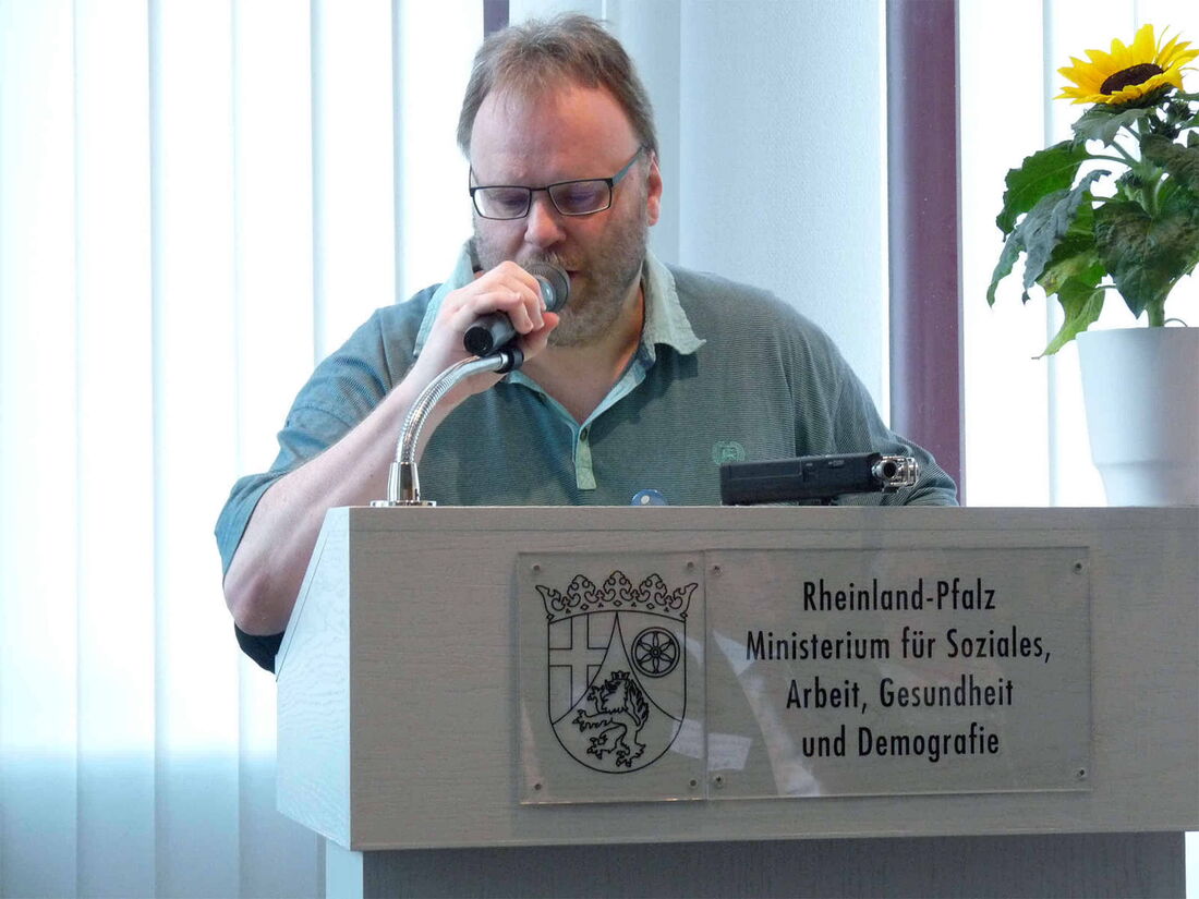 Foto: Hier ergreife ich das Mikrofon am Anfang des ersten Lesungsabschnittes. Ich stehe hinter einem Stehpult. Darauf stehen rechts Sonnenblumen.