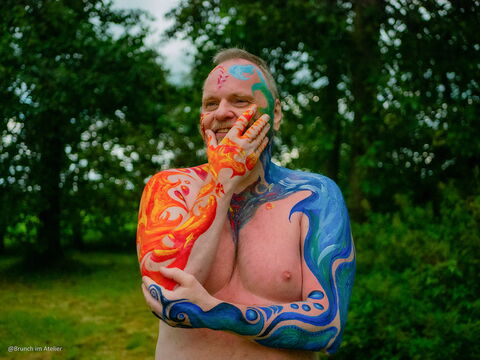 Ich stehe mit bunt bemaltem Oberkörper, Schultern und Gesicht in einem Garten.