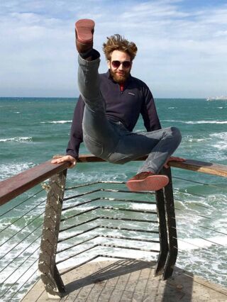 Foto: Benjamin stützt sich mit den Armen am Geländer ab und schmeißt die Beine in die Luft. Im Hintergrund das Meer.