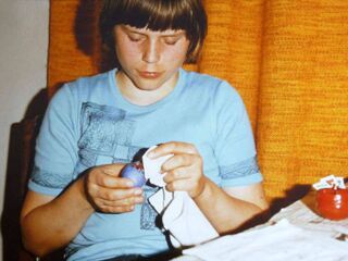 Foto: Ich sitze mit etwa 12 Jahren in einem hellblauen T-Shirt vor orangen Gardinen und bin beim Eierfärben, Ostern um 1980.