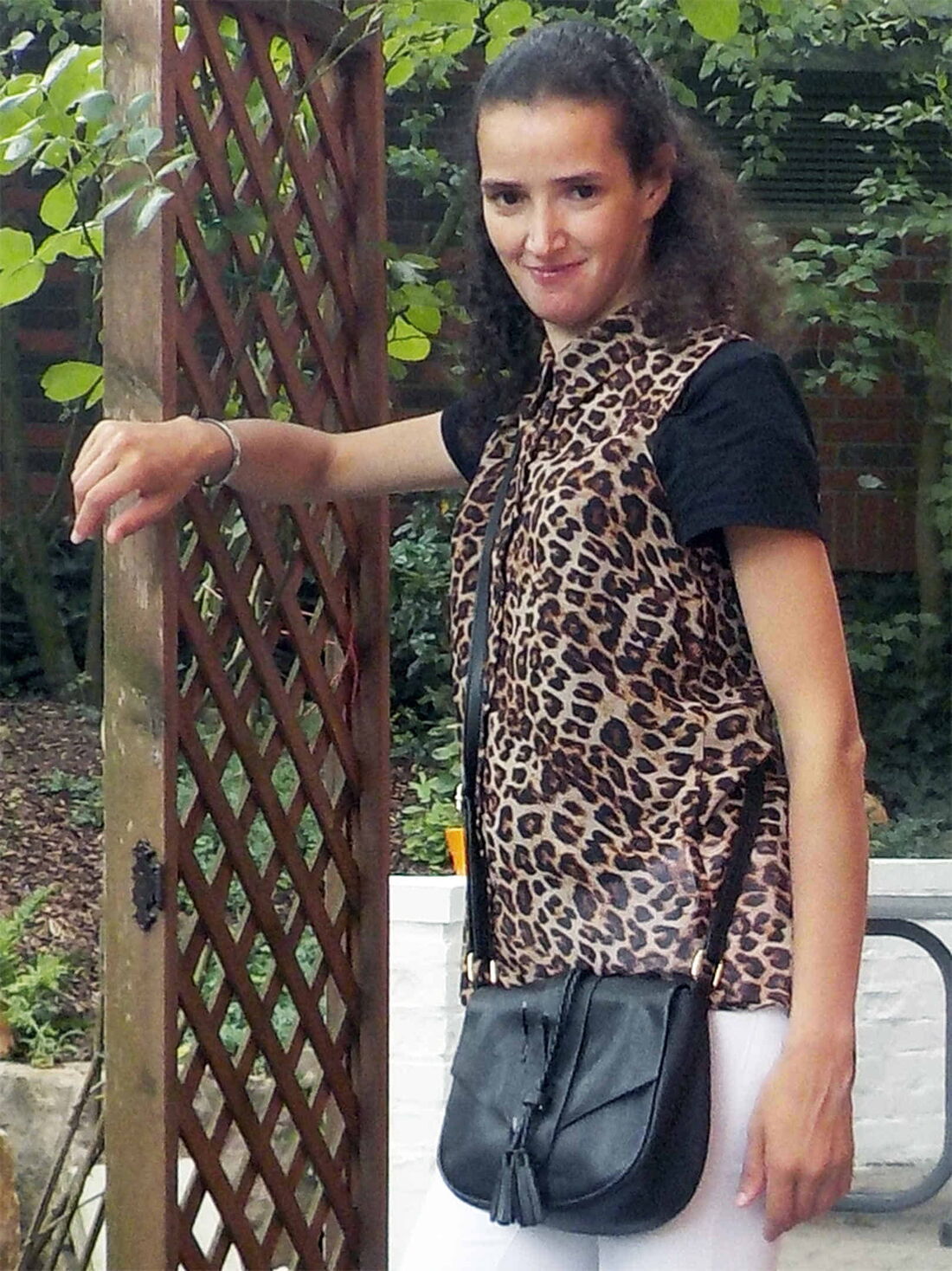 Foto: Fikria mit schwarzem T-Shirt, einer Leopardenweste, einer weißen Hose und schwarzer Handtasche in einer Gartenlaube.