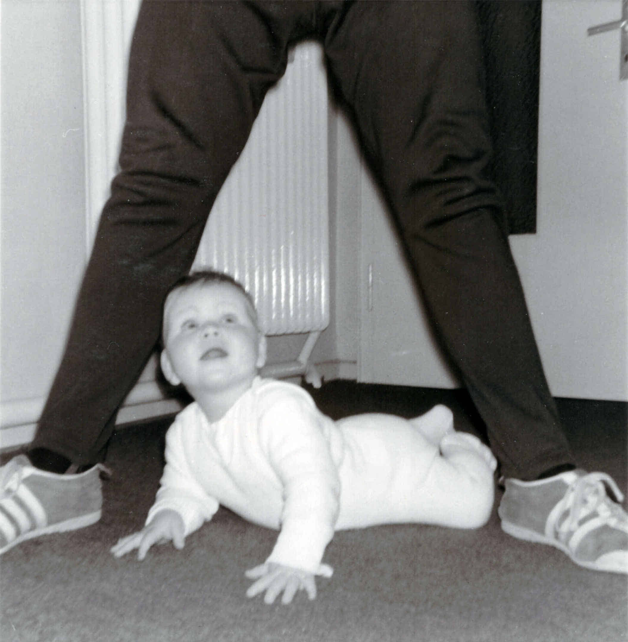 Foto: Ich blicke als Baby auf dem Boden liegend nach oben, während mein Vater mit gespreizten Beinen über mir steht.
